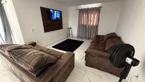 A living room with two brown sofas, a black rug, a standing fan, and a TV mounted on the wall. - Casa Pé na Areia, cozy for guests with good taste. (Praia da Costa Armação dos Búzios)