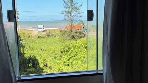 A window with a view of a grassy field, a tree, and a truck. - Casa Pé na Areia, cozy for guests with good taste. (Praia da Costa Armação dos Búzios)