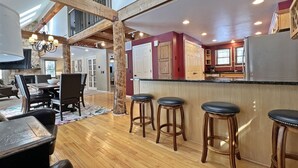 Een ruime woonkamer met een hoge plafond, houten balken, een eettafel met stoelen, een keuken met barstoelen en een trap naar een verdieping erboven.