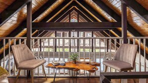 A cozy attic room with wooden beams, two plush chairs, a wooden coffee table with a plant, and a view through a large window. - Spacious 4BR Cabin with a Hot Tub (Munds Park)