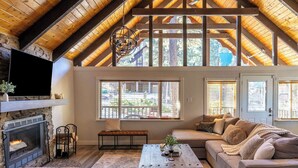 A cozy living room with a stone fireplace, wooden ceiling beams, and large windows. - Spacious 4BR Cabin with a Hot Tub (Munds Park)