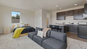 A modern apartment with a large bed, a grey sofa, a kitchen with dark cabinets, and a window providing natural light. - Spacious Pine Retreat with Fire Pit (Flagstaff)