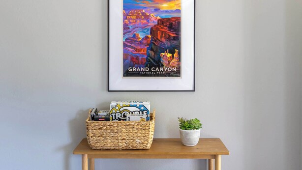 Een ingelijste poster van het Grand Canyon National Park aan een muur, een rieten mand met spullen op een houten tafel eronder, en een potplant.