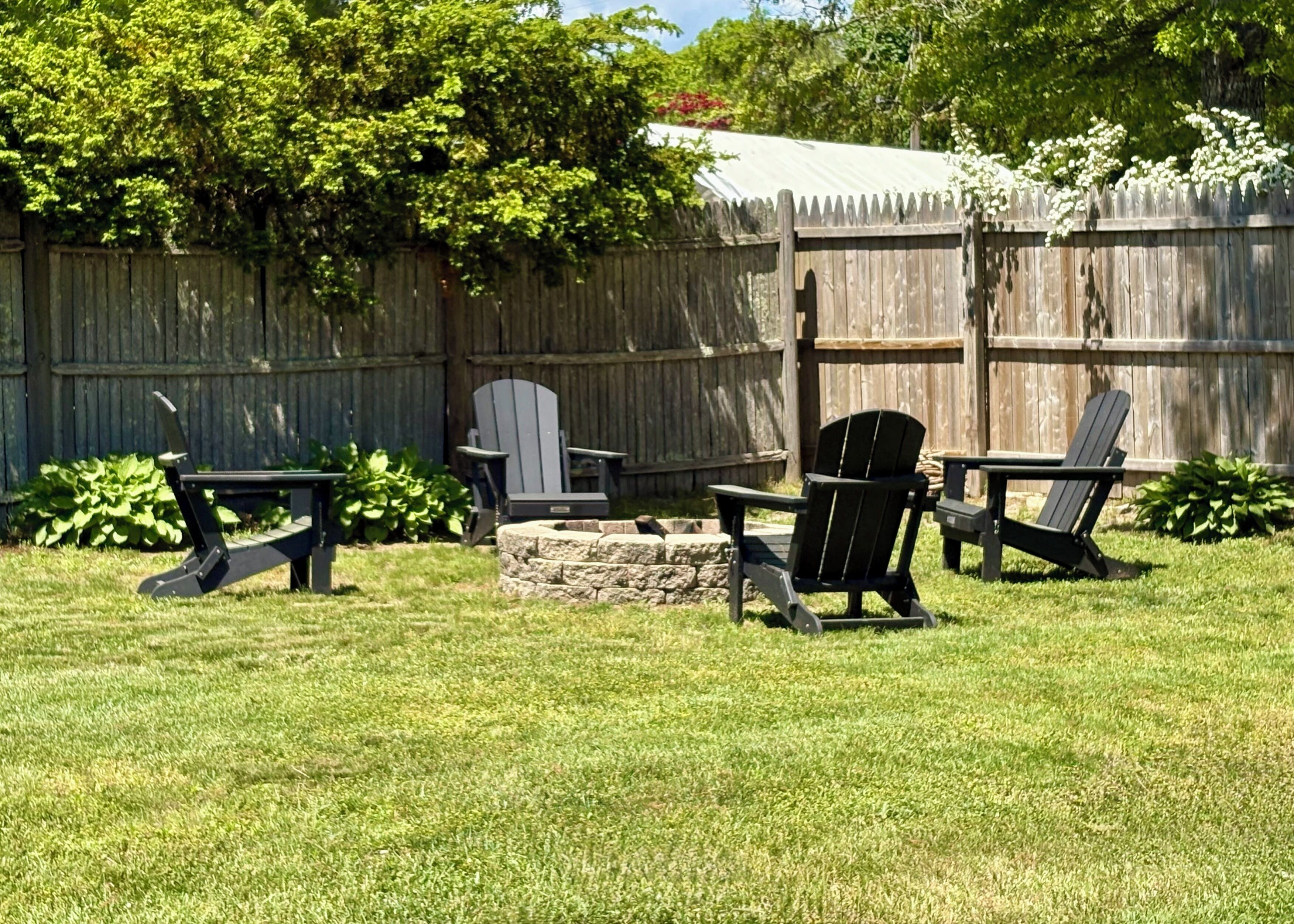A backyard with a stone fire pit, two Adirondack chairs, and a wooden fence.
