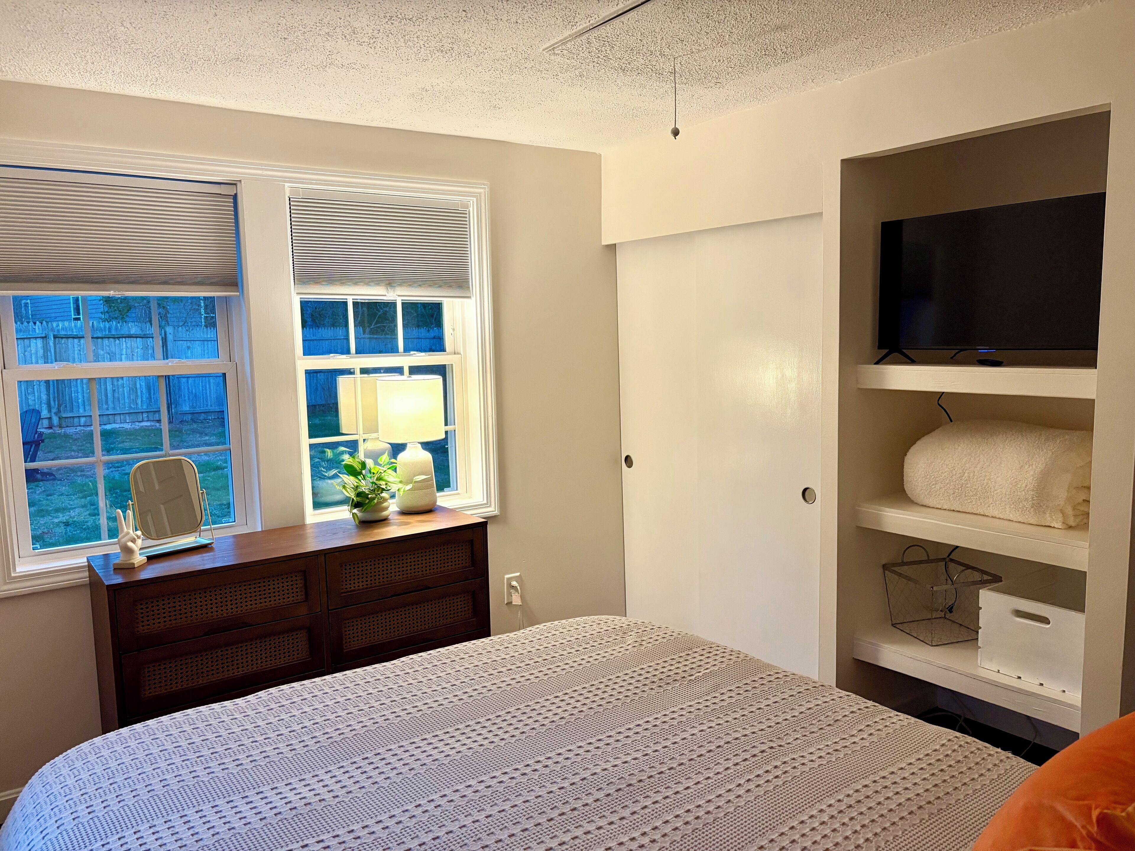 A bedroom with a bed, a dresser, a TV on a shelf, and a window with blinds.
