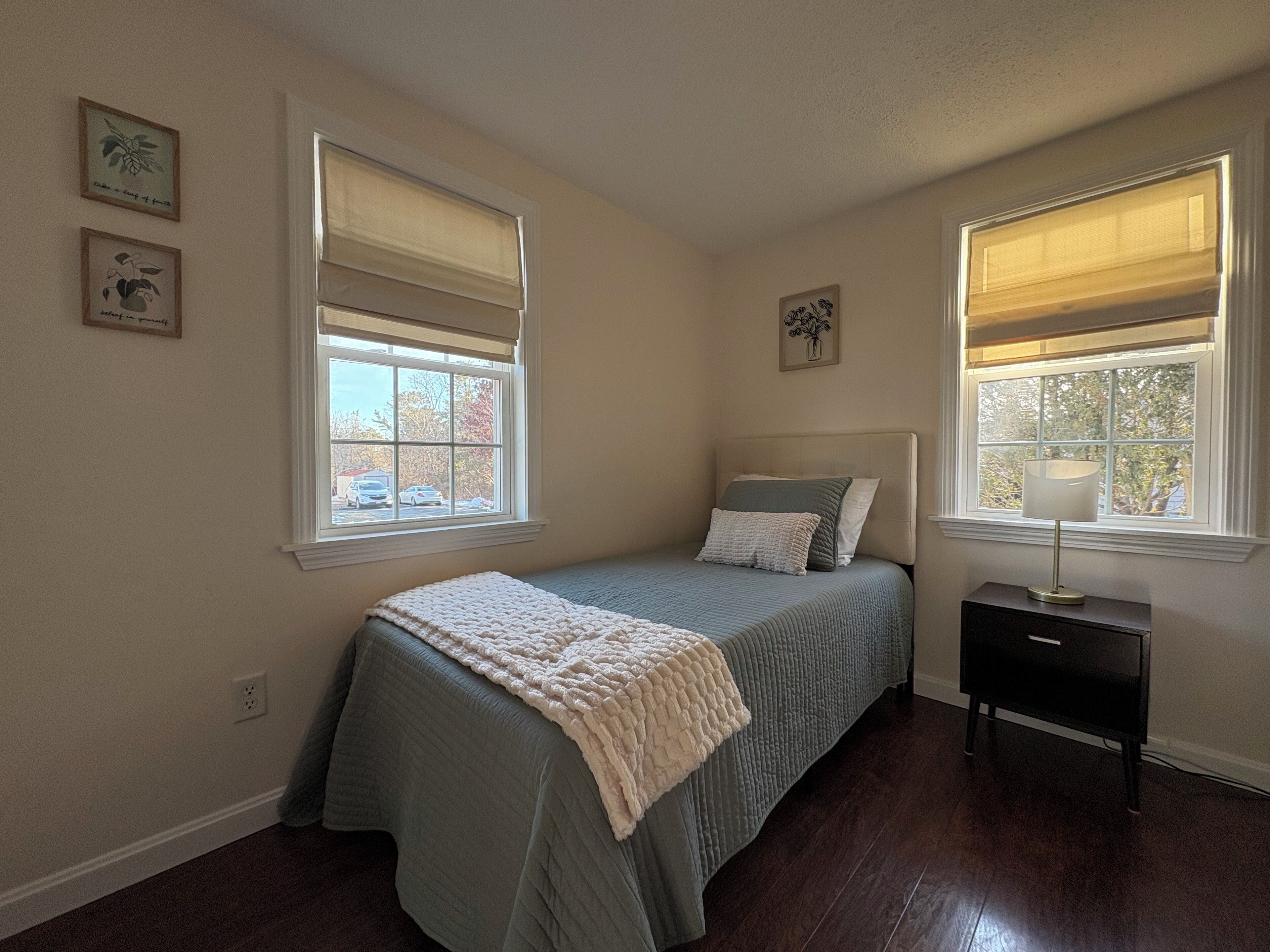 Une chambre à coucher avec un lit, deux fenêtres équipées de stores, une table de chevet et une lampe fixée au mur.