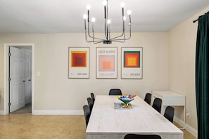 A dining area with a long table, chairs, a chandelier, and three framed artworks on the wall.