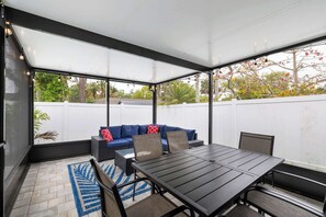 Een overdekt terras met een tafel en stoelen, een bank met kussens, en uitzicht op de achtertuin door grote ramen.