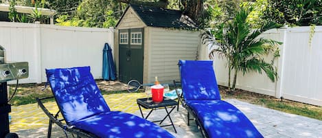 Un patio trasero con dos sillones azules, una parrilla y un cobertizo pequeño.
