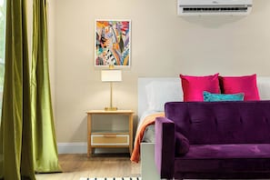 A room with a purple sofa, a small wooden nightstand with a lamp, a colorful abstract painting on the wall, and a window with green curtains.