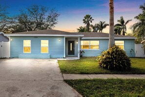 A light blue single-story house with a front door, two windows on the front, and a small front yard with a palm tree.