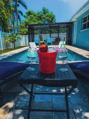 A poolside setting with a table, two chairs, a bottle of champagne, and two wine glasses.