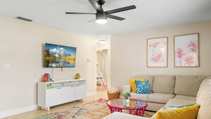 A living room with a sofa, a coffee table, a ceiling fan, a TV mounted on the wall, and a staircase in the background.