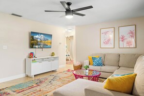 A living room with a sofa, a coffee table, a ceiling fan, a TV mounted on the wall, and a staircase in the background.