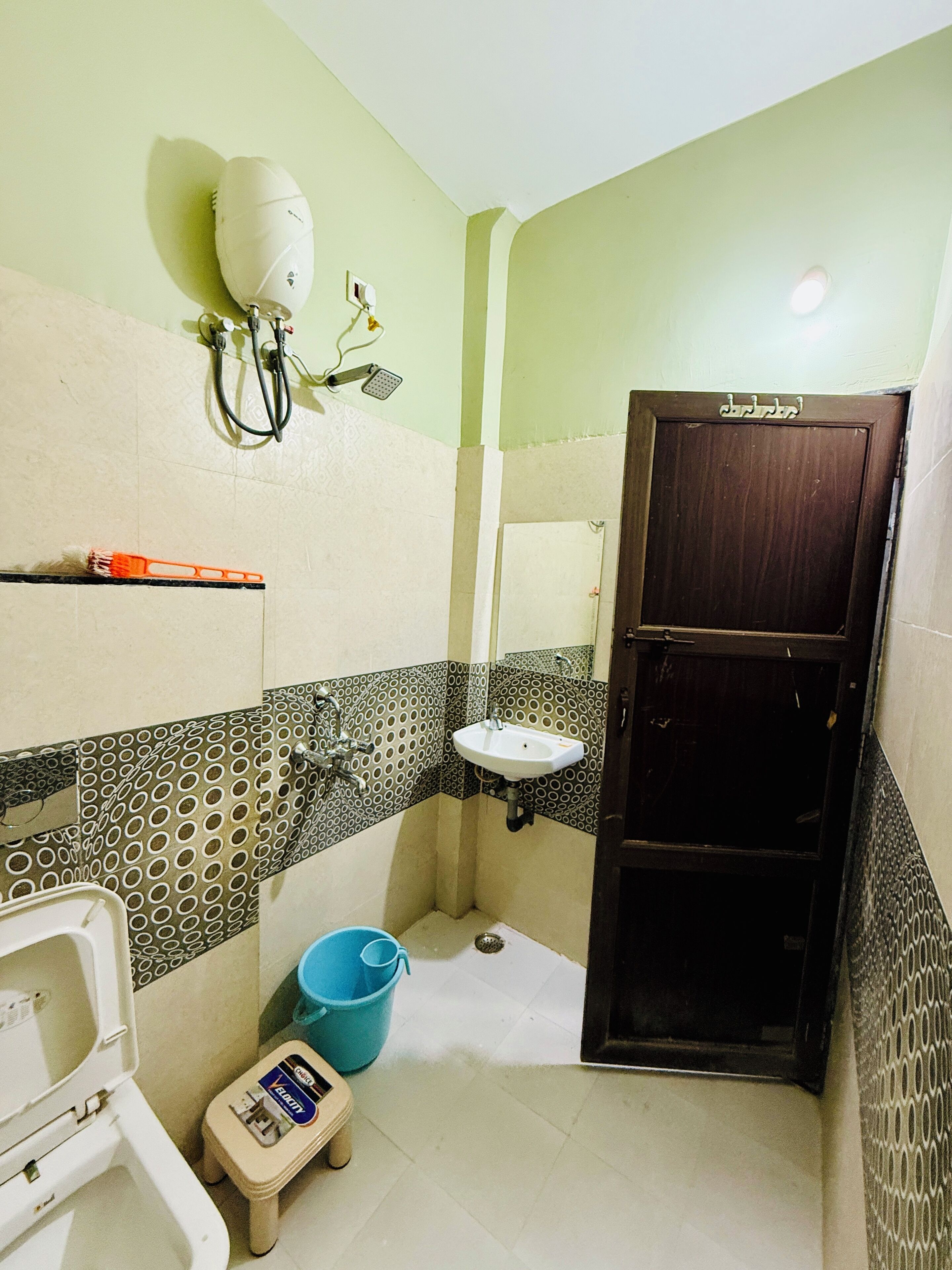 A small bathroom with a toilet, sink, and shower. There is a blue bucket and a small stool.