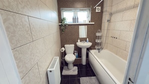 A compact bathroom with a toilet, sink, and bathtub. There is a window above the sink and a radiator on the floor. - Cozy House Sleeps 2 With Free Parking (Birmingham)