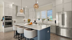 A modern kitchen with white cabinetry, stainless steel appliances, a central island with bar stools, pendant lighting, and a view of the outside through large windows. - Villa Capri · 2025 Luxury Build · Steps to Beach (Fort Myers Beach)