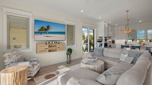 A spacious living room with a large sectional sofa, a coffee table, and a television mounted on the wall. There is a kitchen area visible in the background. - Villa Capri · 2025 Luxury Build · Steps to Beach (Fort Myers Beach)