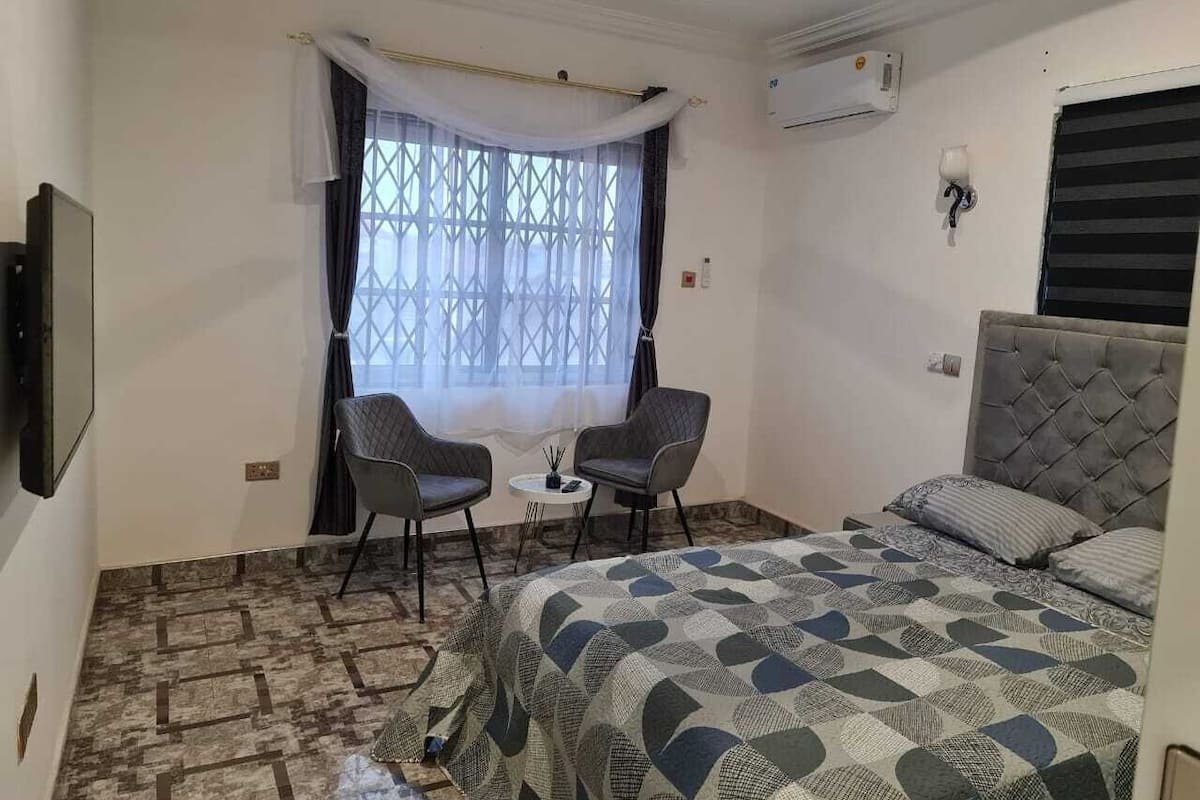 Une chambre d’hôtel avec un lit, deux chaises, une petite table et une fenêtre avec des rideaux.