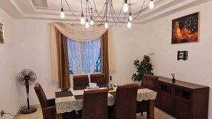 A dining area with a round table, chairs, a fan, a cabinet, and a window with curtains. - King George Lodge  (Tema)
