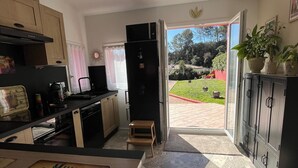 A compact kitchen with a small dining area, a refrigerator, and a view of a garden through an open door. - WELCOME TO "LOU PARADOU" NEW HOUSE FOR 4 PEOPLE IN PROVENCE VERTE (Méounes-lès-Montrieux)