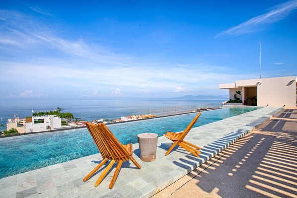 An infinity pool with clear blue water, two wooden lounge chairs, a stone pedestal, and a view of the ocean and distant buildings.