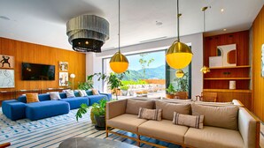 Una hall d'albergo moderna con un grande divano, poltrone blu e beige, un tavolino da caffè nero e un televisore montato a parete.