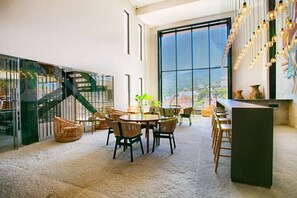 Amplio espacio interior con techo alto, ventanas grandes y una escalera que sube a un nivel superior. Varias zonas de estar con mobiliario de mimbre y una barra.