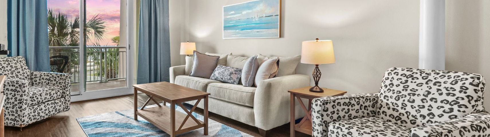 Sala de estar com sofá, poltrona e mesa de centro, varanda com cortinas e um quadro na parede.