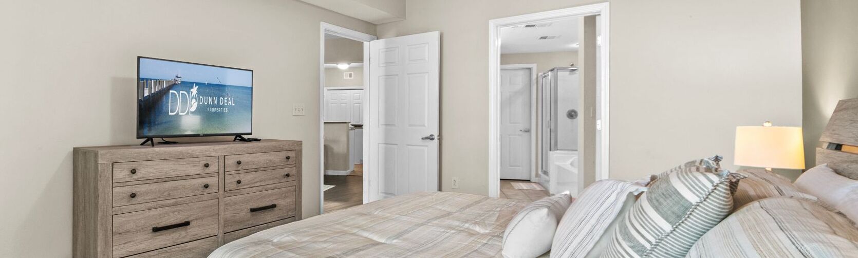 A bedroom with a bed, a wooden dresser, a television, and a door leading to a bathroom.