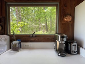 A kitchen with a window overlooking a forest, a coffee maker, a toaster, and a jar of sugar.
