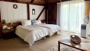 Ein Schlafzimmer mit Holzboden, einem Bett mit weißer Bettwäsche, einem Nachttisch mit Lampe, einem Fenster mit durchsichtigen Vorhängen und einer Wand-Klimaanlage.