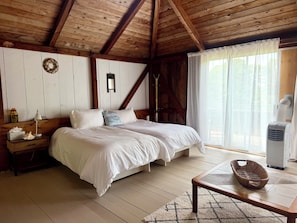 A bedroom with a wooden ceiling, a bed with white bedding, a bedside table with a lamp, a window with sheer curtains, and a wall-mounted air conditioner.