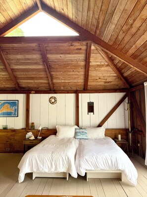 A bedroom with a wooden ceiling, a bed with white bedding, a nightstand, a map on the wall, and a small table with a lamp. - Cozy 3BR Cottage with Lambert's Cove Beach access (West Tisbury)