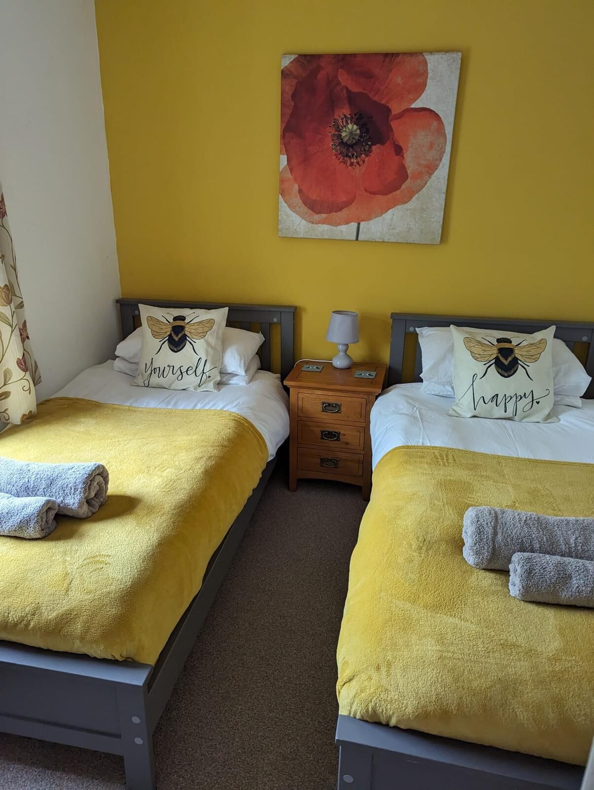 A room with two beds, each with a yellow blanket and a pillow featuring a bee design. There is a nightstand with a lamp between the beds, and a large flower painting on the wall.