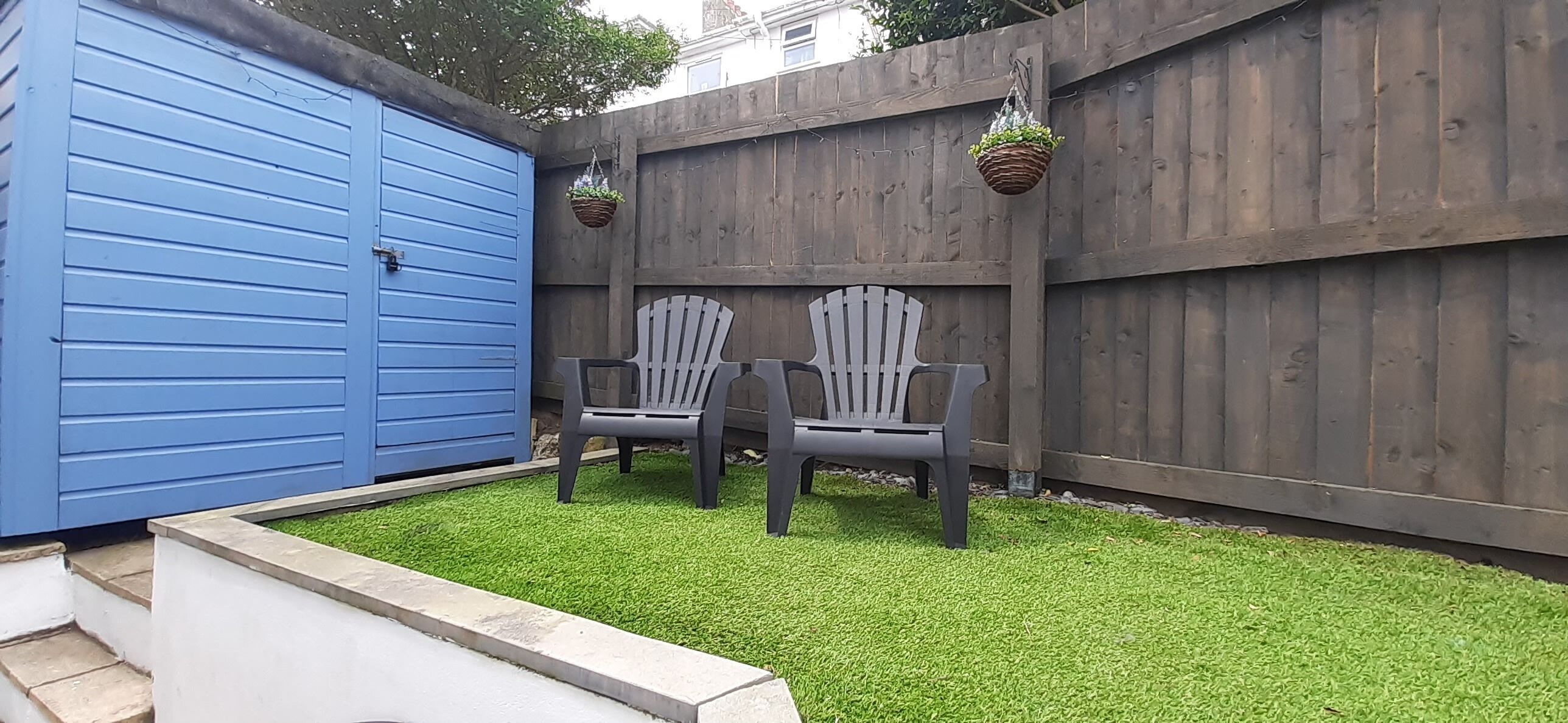 Un jardin avec une pelouse artificielle, deux fauteuils Adirondack, une porte de garage bleue et une clôture en bois.