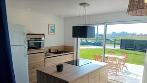A modern kitchen with a built-in oven, a sink, and a large island. There is a dining area with a table and chairs outside, and a view of a green landscape. - Recent house facing the dunes and Grève Blanche beach, at the foot of the GR34. (Plouguerneau)