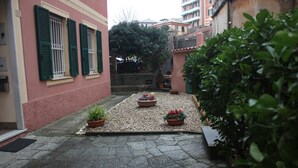 A courtyard with potted plants, a stone pathway, and a building with green shutters. - Tre Cuori - Amy. (Genova)