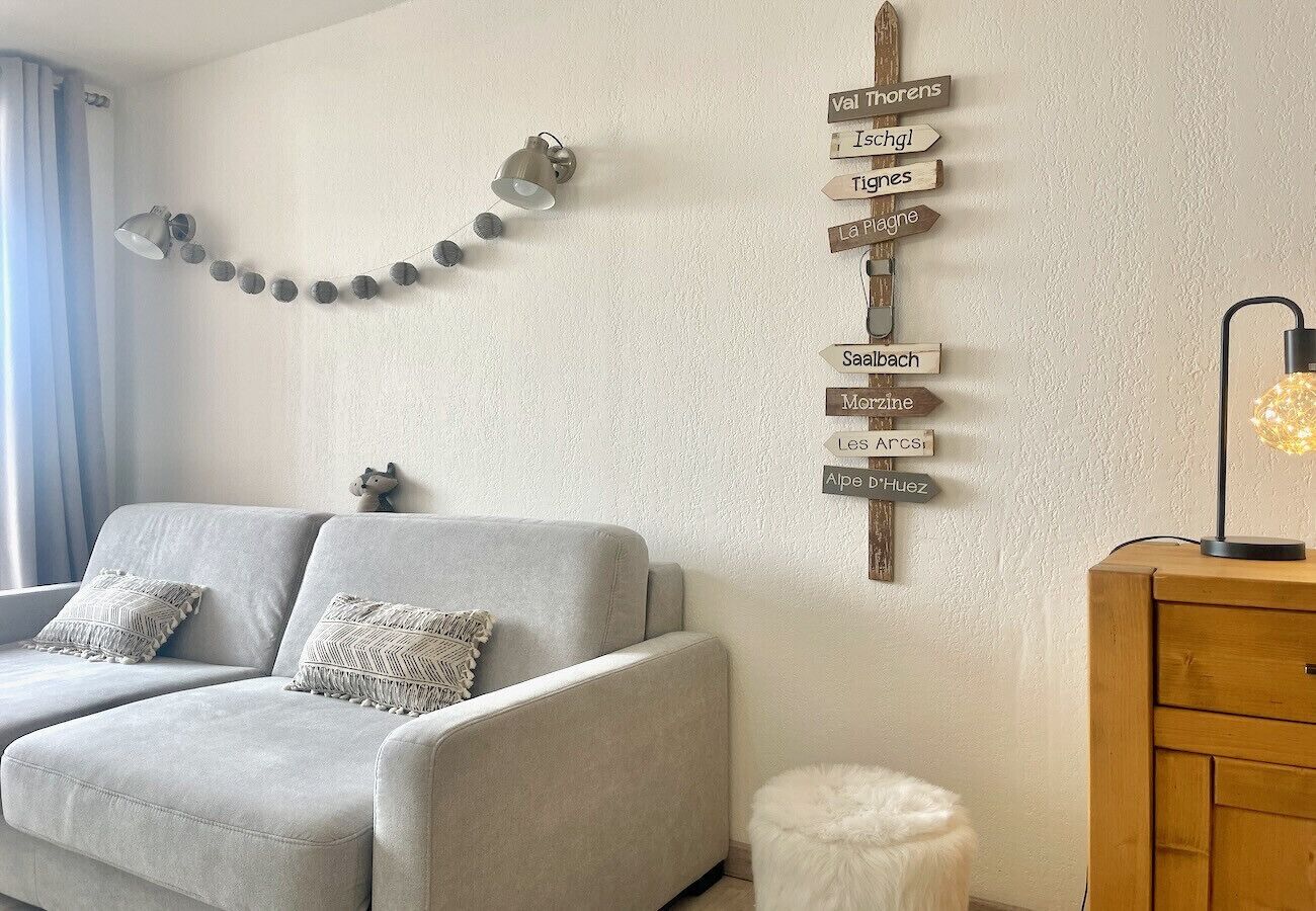 A living room with a grey sofa, two patterned cushions, a string of spherical lights, a wall-mounted sign with various names, and a lamp on a wooden side table.