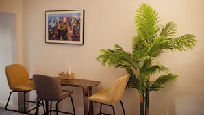 A dining area with a table and two chairs, a framed cityscape picture on the wall, and a large potted plant.