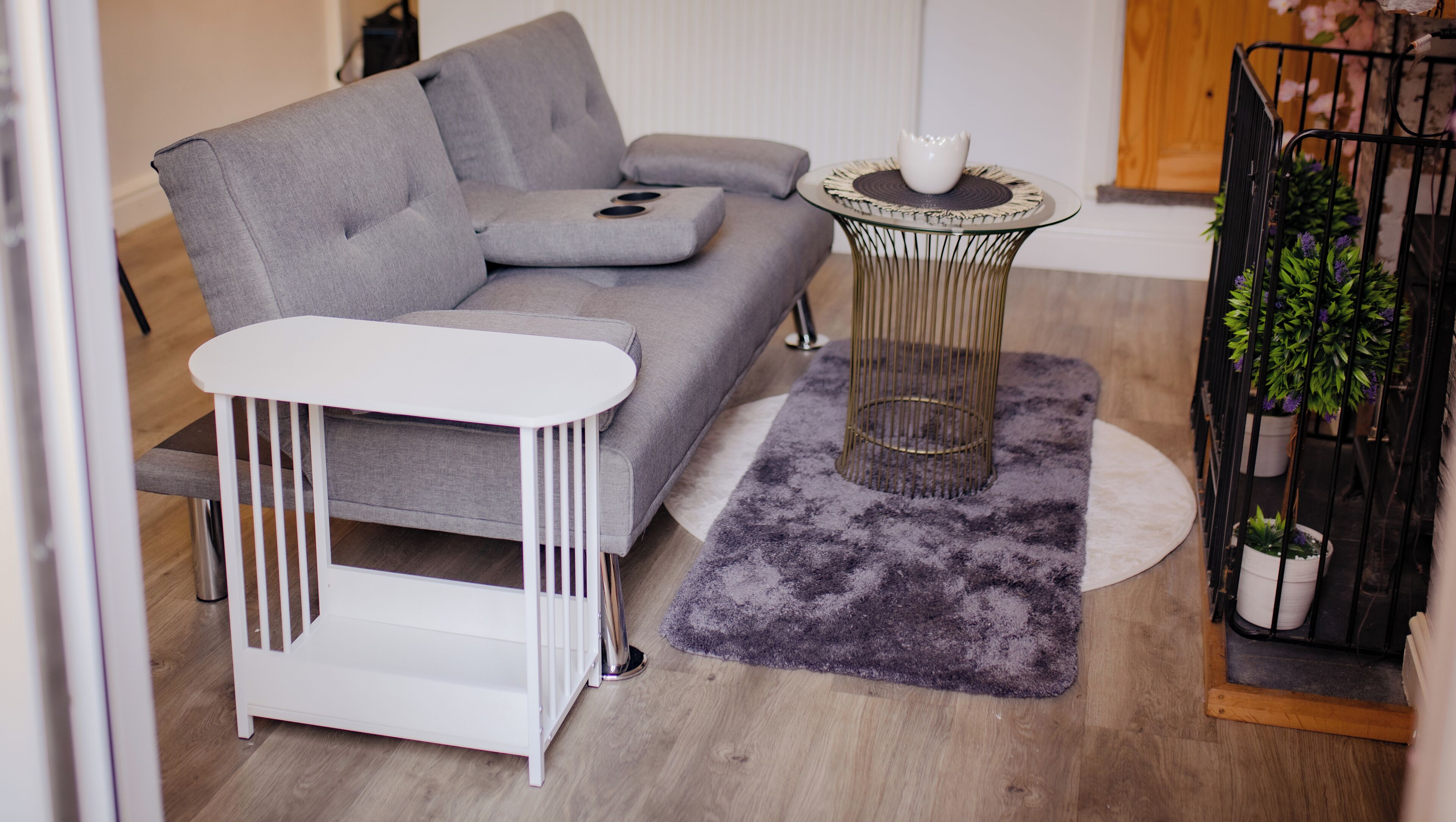 Een moderne woonkamer met een grijze bank, een witte bijzettafel, een glazen salontafel met een witte schaal, een paars vloerkleed en een zwarte metalen plantenstandaard met groene planten.