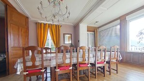 Une salle à manger avec une table et des chaises en bois, un lustre et de grandes fenêtres avec des rideaux.