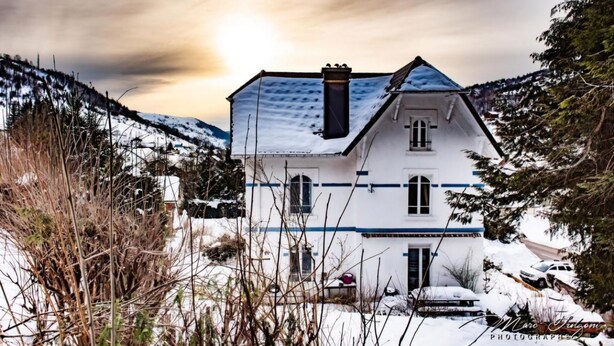 Une maison blanche à deux étages, surmontée d’un toit bleu, entourée d’arbres et de collines couverts de neige.