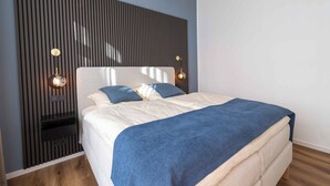 Un lit bien fait, recouvert d’un plaid bleu, avec une tête de lit à lattes verticales et deux lampes de lecture fixées au mur.