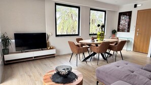 A modern living room with a TV, dining table, and chairs. There are windows with a view of palm trees. - Solsalt 1min Walk to Beach 1min From Everything (Platanias)