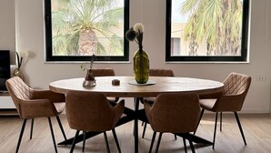 A dining area with a round table, six chairs, and two windows with views of palm trees. - Solsalt 1min Walk to Beach 1min From Everything (Platanias)