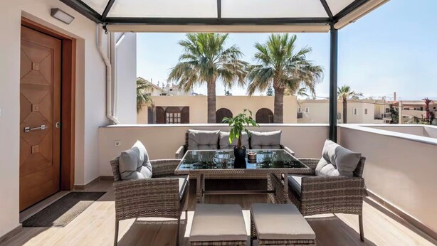 An outdoor balcony with a glass table and wicker furniture, overlooking palm trees and buildings. - Solsalt 1min Walk to Beach 1min From Everything (Platanias)