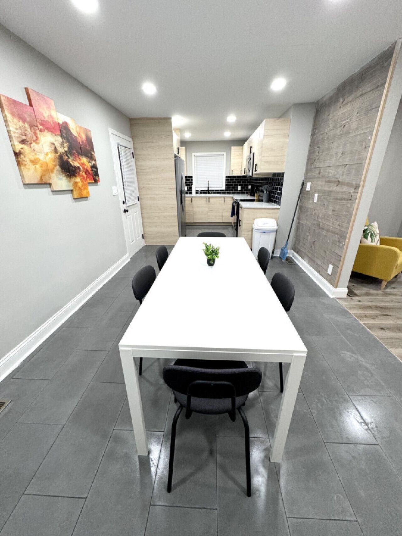 A modern dining area with a white table and black chairs, a kitchen in the background, and a gray floor.