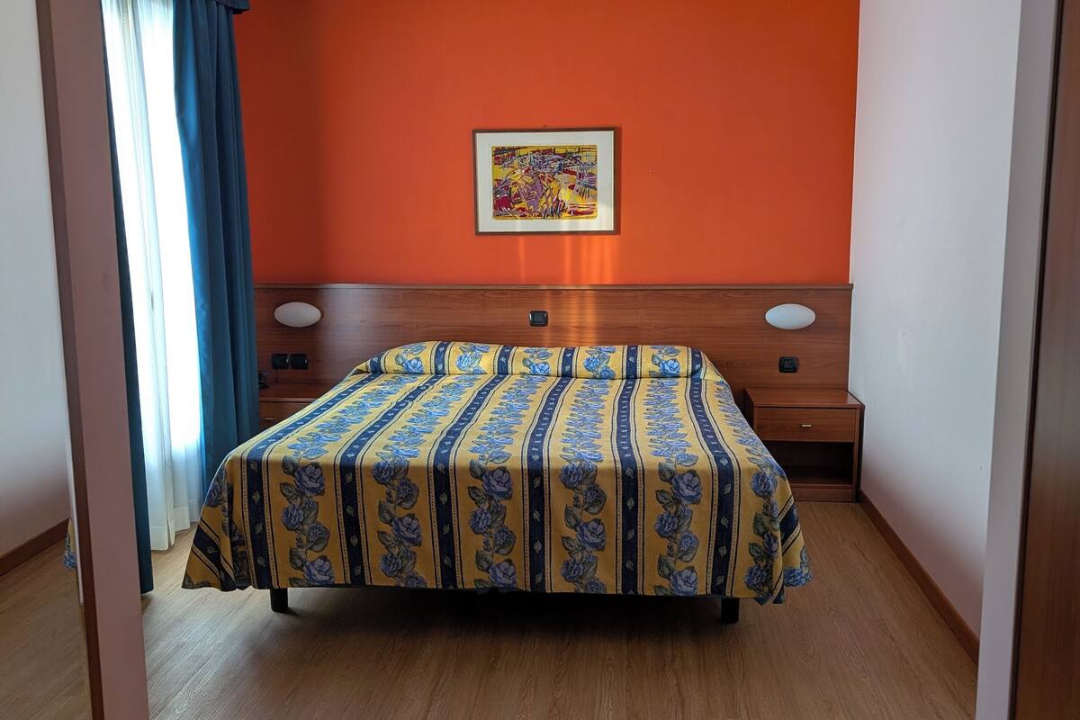Habitación de hotel con cama, cabecera de madera, un cuadro enmarcado en la pared y una ventana con cortinas.