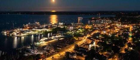Un pueblo costero de noche con un puerto deportivo, edificios y una torre destacada.